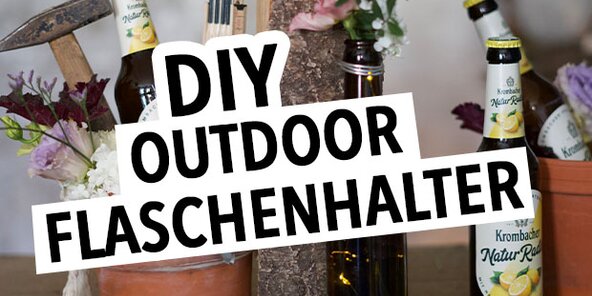 Zwei Flaschen Krombacher Natur-Radler stehen in einem Blumentopf und einem Holzblock. Ein Holzhammer und Blumen sind ebenfalls sichtbar. Der Text 'DIY OUTDOOR FLASCHENHALTER' ist prominent in Schwarz auf Weiß.