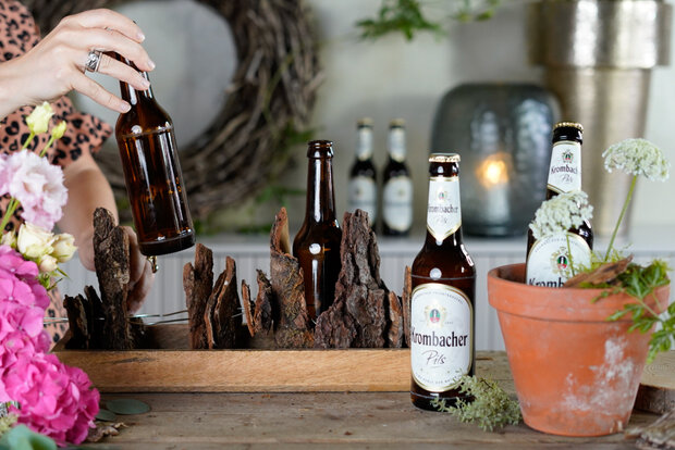 Aufgefädelte Rindenstücke werden auf ein Holztablett gelegt und mit brauen Bierflaschen dekoriert.