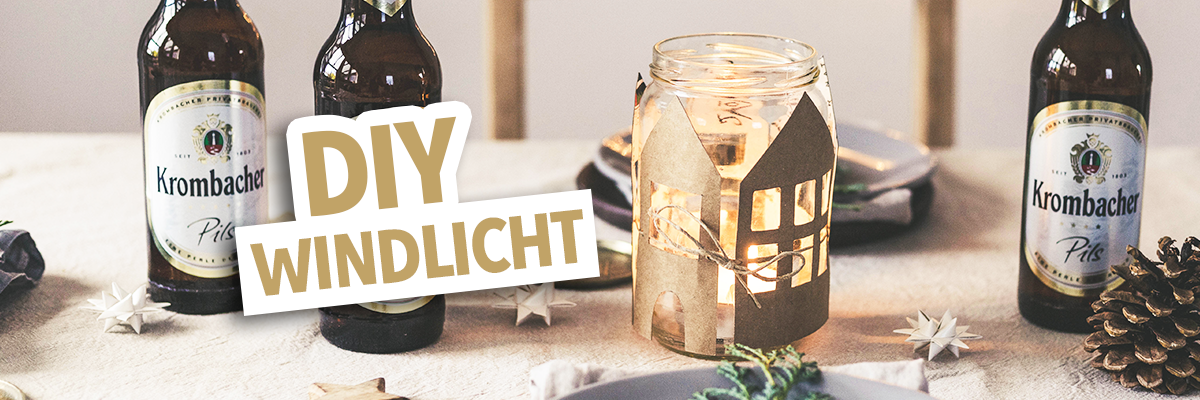 Zwei Flaschen Krombacher Pils stehen auf einem Tisch neben einem DIY-Windlicht in Form eines Hauses. Der Text 'DIY WINDLICHT' ist prominent in der Mitte des Bildes platziert.
