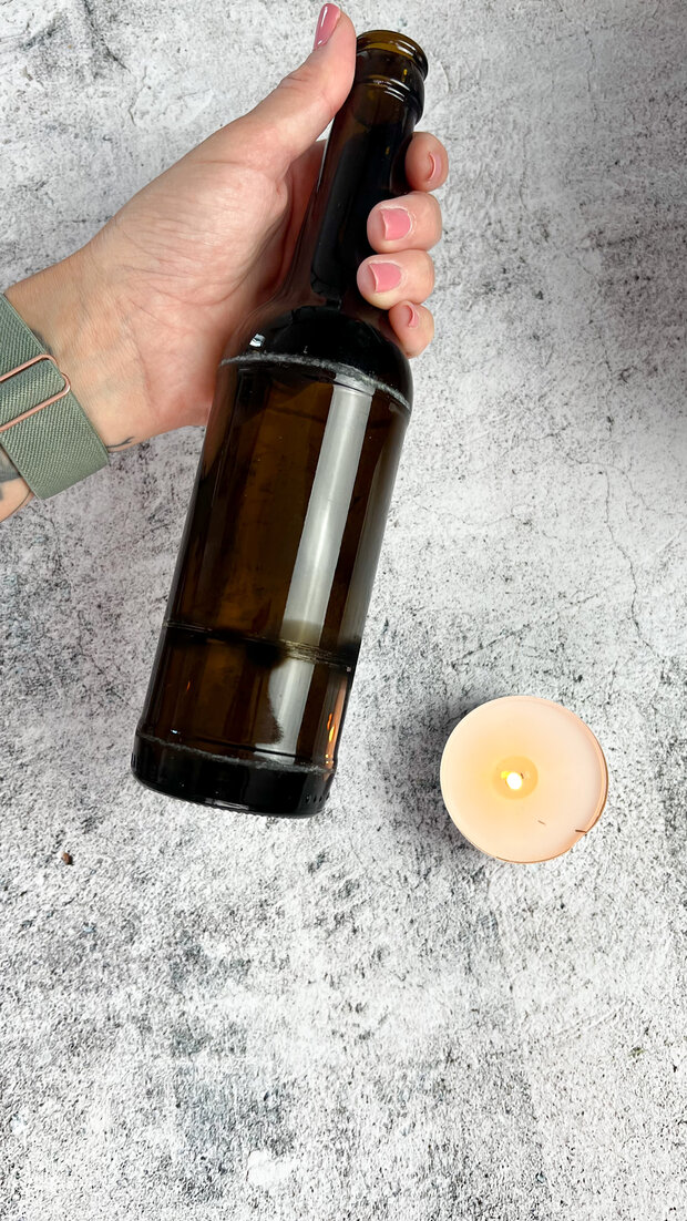 Eine Hand hält eine braune Glasflasche mit schwarzem Deckel. Neben der Flasche steht eine brennende Kerze in einem weißen Teelichtbehälter.