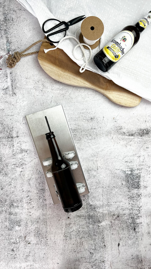 Ein schwarzes Flaschenöffnergerät mit einer dunklen Flasche 'Grünhoffs' daneben. Im Hintergrund sind eine Holzplatte, eine Schere und eine Rolle Kraftpapier sichtbar.