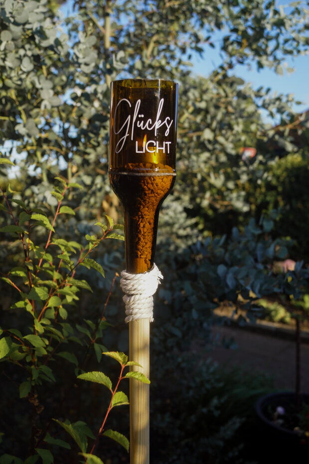 Ein Glas mit der Aufschrift 'Glücks LICHT' steht auf einem Holzpfahl, umgeben von grünen Pflanzen. Das Glas hat eine braune Farbe und ist mit einem weißen Seil am Pfahl befestigt.