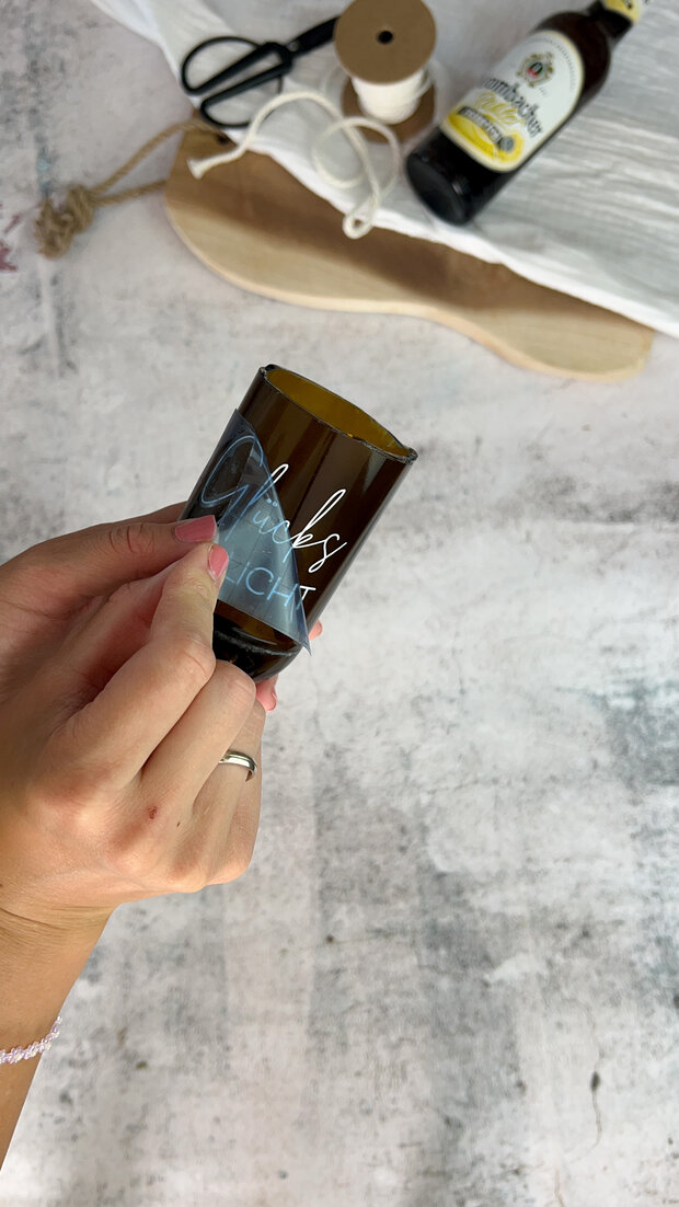 Eine Person hält ein braunes Glas mit der Aufschrift 'Glücksbringer'. Im Hintergrund sind eine Schnur, ein Schuh und eine Flasche Bier sichtbar.