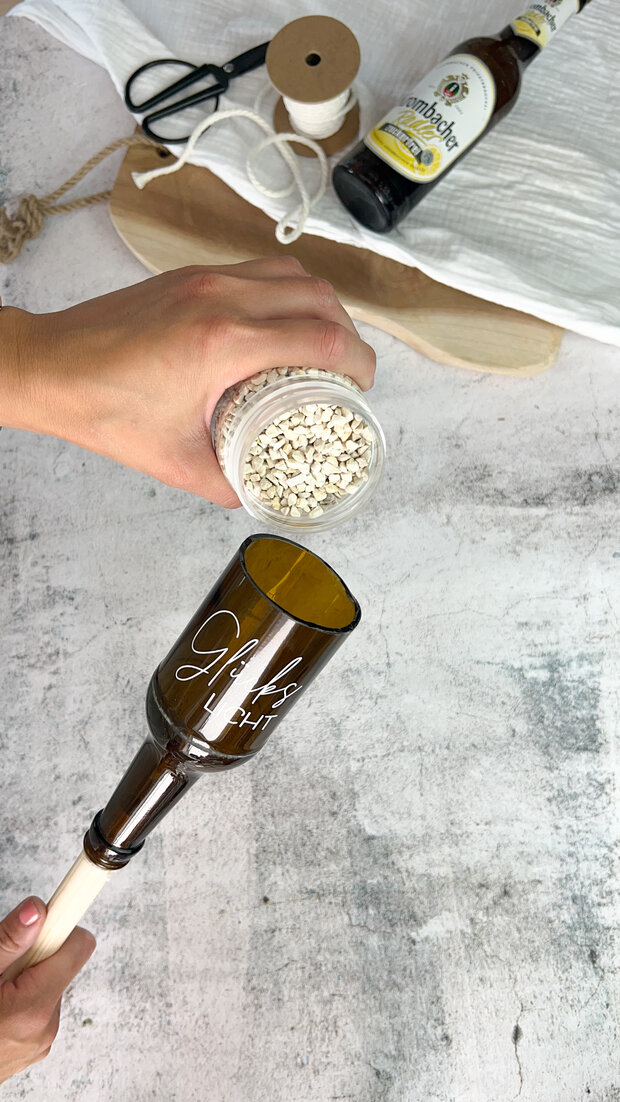 Eine Hand hält ein Glasgefäß mit weißen Bohnen über ein umgedrehtes, graviertes Flaschengefäß mit dem Namen 'Jules'. Im Hintergrund steht eine Flasche Bier und eine Schere.