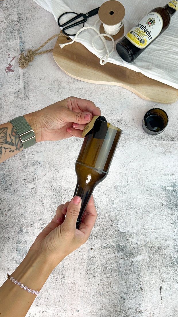 Eine Person mit tätowierten Armen hält eine braune Glasflasche, die oben abgeschnitten ist. Auf dem Tisch liegen eine Schere, eine Holzplatte, ein Garnrollen und eine Flasche 'Hefeweizen' von 'Paulaner'.