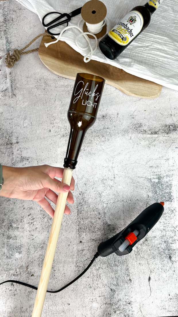 Eine Person hält einen Holzstab, an dessen Ende eine braune Glasflasche mit der Aufschrift 'Glücks LIGHT' befestigt ist. Im Hintergrund sind eine Schere, ein Garnrollenhalter und eine Flasche Bier sichtbar.