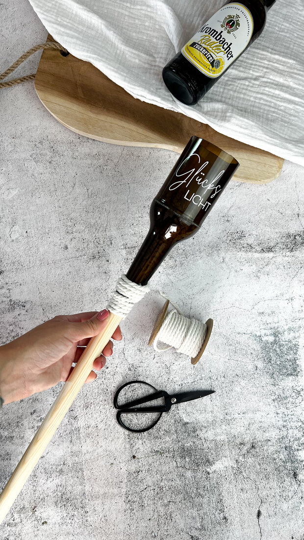 Eine braune Glasflasche mit der Aufschrift 'Gäste Licht' ist an einem Holzstab befestigt. Daneben liegt eine Rolle mit weißem Garn und eine Schere.