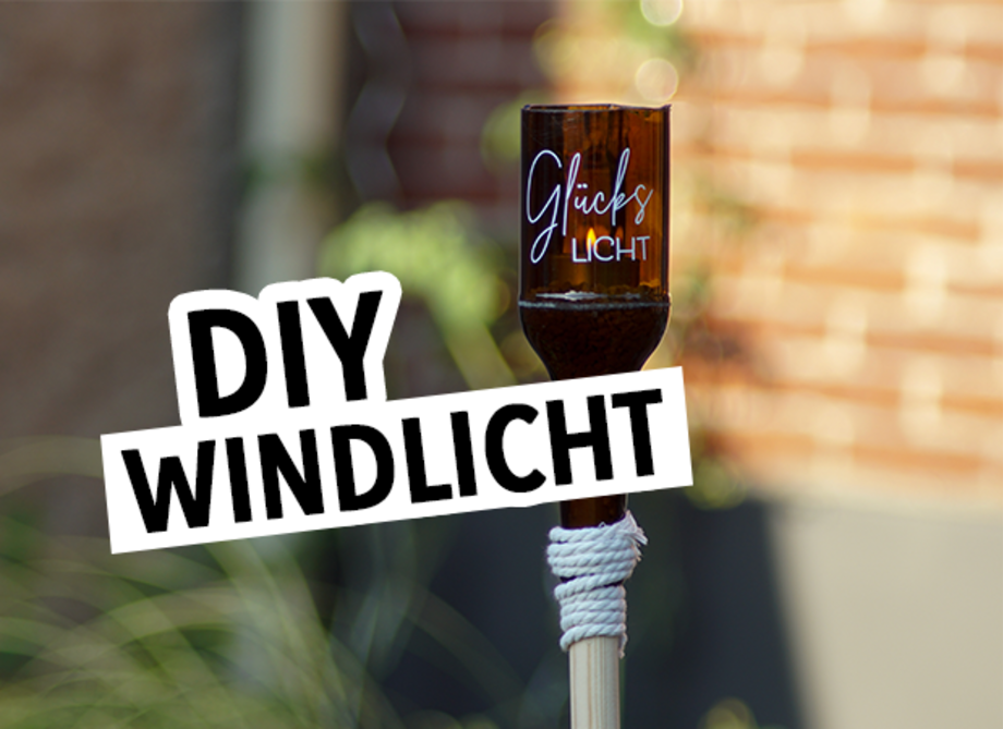 Ein braunes Glaswindlicht mit der Aufschrift 'Glückslicht' steht auf einem Holzstab. Der Hintergrund zeigt eine unscharfe, grüne Pflanzenlandschaft. Der Text 'DIY WINDLICHT' ist in großen, schwarzen Buchstaben im Vordergrund.