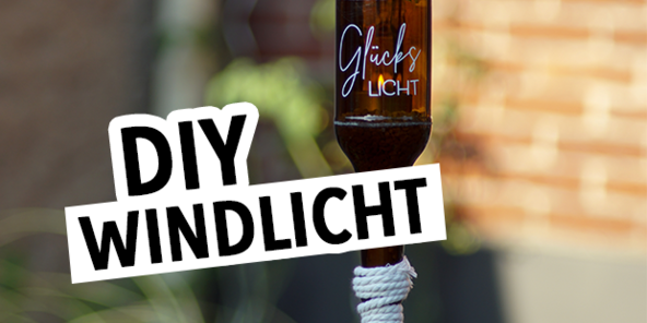 Ein braunes Glaswindlicht mit der Aufschrift 'Glückslicht' steht auf einem Holzstab. Der Hintergrund zeigt eine unscharfe, grüne Pflanzenlandschaft. Der Text 'DIY WINDLICHT' ist in großen, schwarzen Buchstaben im Vordergrund.