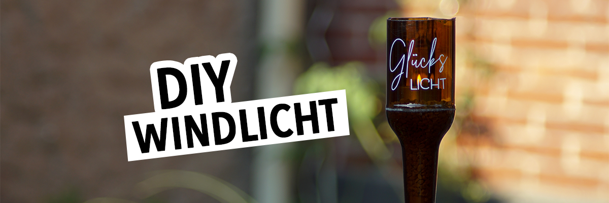 Ein braunes Glaswindlicht mit der Aufschrift 'Glückslicht' steht auf einem Holzstab. Der Hintergrund zeigt eine unscharfe, grüne Pflanzenlandschaft. Der Text 'DIY WINDLICHT' ist in großen, schwarzen Buchstaben im Vordergrund.