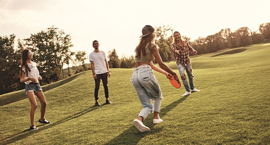 Vier Personen spielen fröhlich zusammen im Kreis Frisbee auf einer Wiese in der Abendsonne.