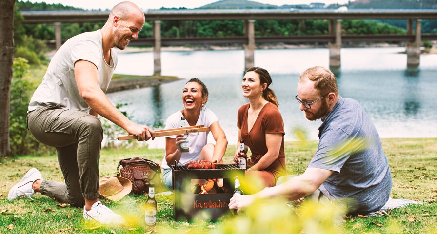 Vier Personen um einen Grill an einem See mit Krombacher Pils Getränken.