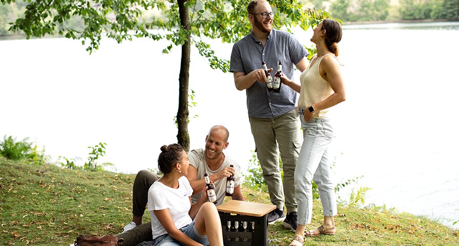 Vier Menschen sitzen und stehen zusammen und stoßen auf einer Krombacher Pils auf einer Wiese an einem See an.