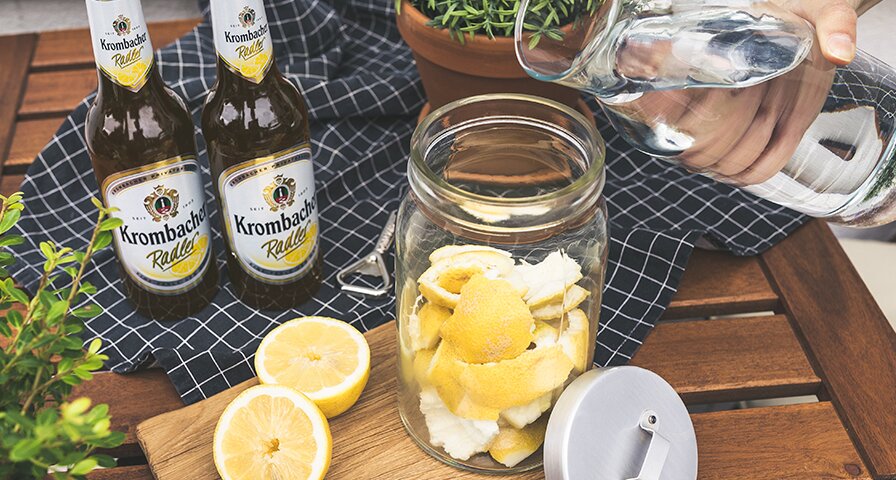 In ein Glas mit Zitronenschalen wird stilles Wasser reingeschüttet. Zwei Krombacher Radlerflaschen stehen auf dem Tisch.
