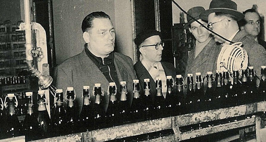 Drei Personen stehen an einer Abfüllanlage, während Flaschen mit Krombacher Bier über ein Förderband laufen.