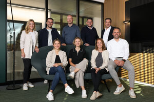 Eine Gruppe von acht Personen sitzt und steht auf einem großen, grünen Sessel in einem modernen Raum. Die Personen tragen legere Kleidung und weiße Schuhe. 