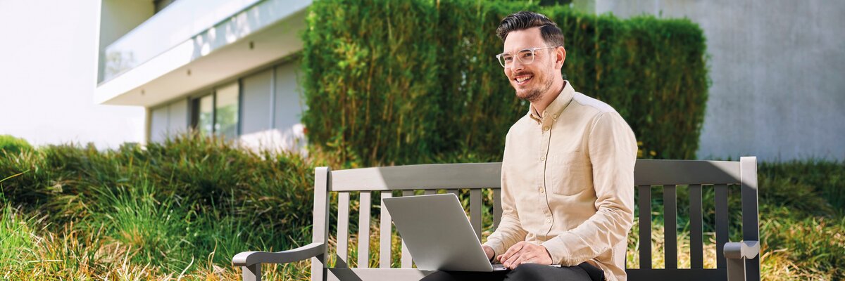 Mann sitzt auf einer Bank im Freien und arbeitet an einem Laptop. Im Hintergrund sind moderne Gebäude und grüne Sträucher sichtbar.