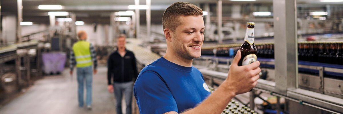 Mann hält eine Flasche Bier in der Hand. Im Hintergrund sind weitere Personen und Flaschen auf einem Förderband zu sehen.