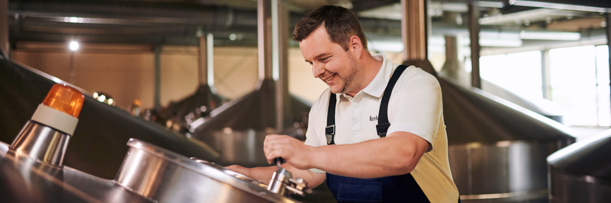 Ein Mann in einem weißen Polohemd und blauen Latzhosen steht an einem großen, runden Behälter in einer Brauerei. Im Hintergrund sind mehrere ähnliche Behälter sichtbar.