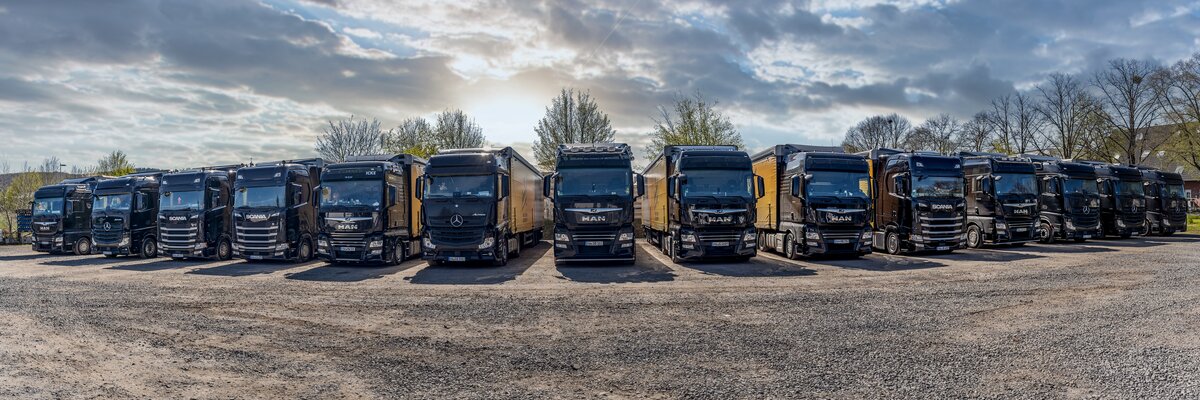 Eine Reihe von 12 schwarzen Lastwagen steht in einer geraden Linie auf einem Schotterplatz. 