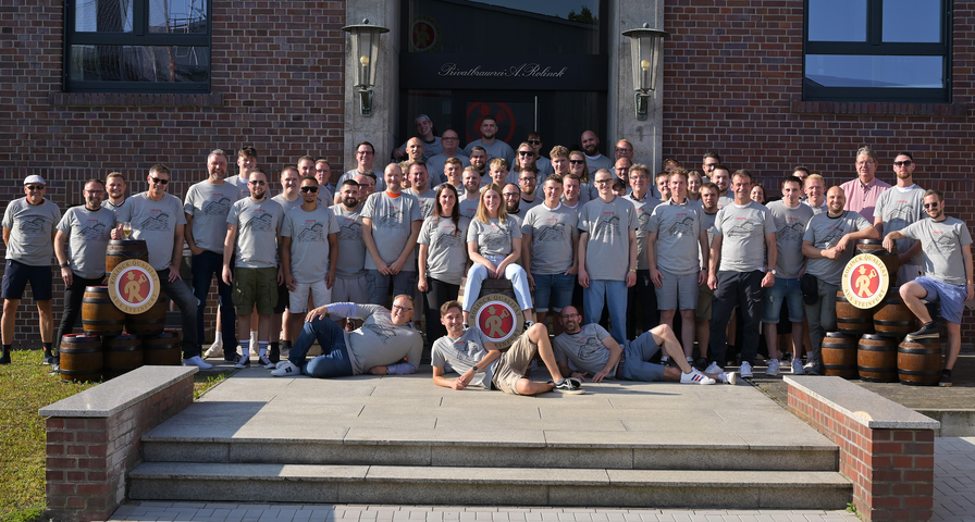 Gruppensfoto von etwa 40 Personen in grauen T-Shirts, die auf einer Treppe vor einem Backsteingebäude stehen. Einige Personen sitzen auf der Treppe oder auf Holzfässern.