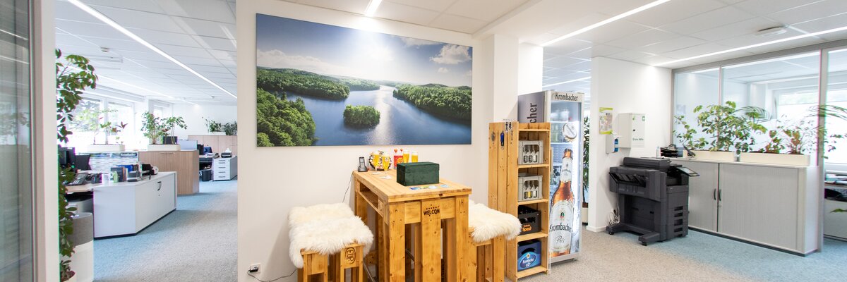 Ein moderner Büroraum mit einem Holztisch und zwei Hockern. Im Hintergrund sind Büromöbel und Pflanzen sichtbar.