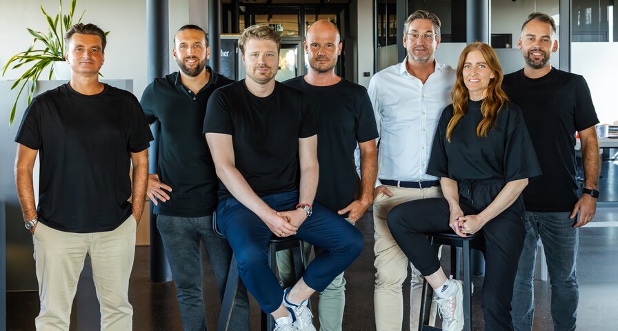 Sechs Personen in schwarzen T-Shirts stehen und sitzen in einer modernen Umgebung. Eine Person trägt eine weiße Hose und Sneakers.