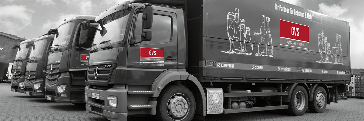 Fünf Lkw der Marke Mercedes-Benz stehen auf einem gepflasterten Parkplatz. Die Lkw sind schwarz-weiß, mit rotem Logo 'GVS' und weiteren grafischen Elementen auf der Seite.