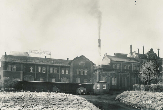Historische Krombacher Brauerei mit mehreren Fenstern und Schornsteinen, aus dem Rauch aufsteigt. Im Vordergrund eine Wiese.