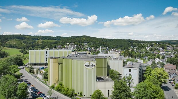 Luftaufnahme der Krombacher Brauerei mit grüner und gelber Fassadenfarbe, umgeben von Bäumen und Wohngebieten.