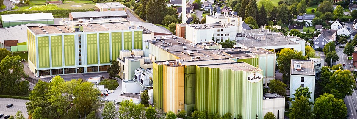 Luftaufnahme der Krombacher Brauerei mit grüner und gelber Fassadenfarbe, umgeben von Bäumen und Wohngebieten.
