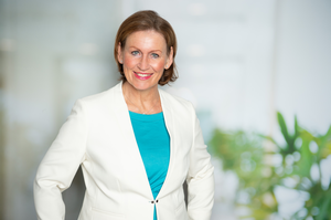 Eine Frau trägt kurze hellbraune Haare und einen weißen Blazer. Sie lächelt in die Kamera.