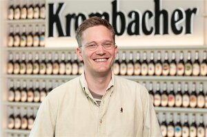 Mann mit blonden Haaren und Brille trägt ein beiges Hemd. Er lächelt in die Kamera.