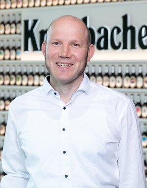 Mann mit kurzen hellen Haaren und einem Dreitagebart, trägt ein weißes Hemd und lächelt in die Kamera.