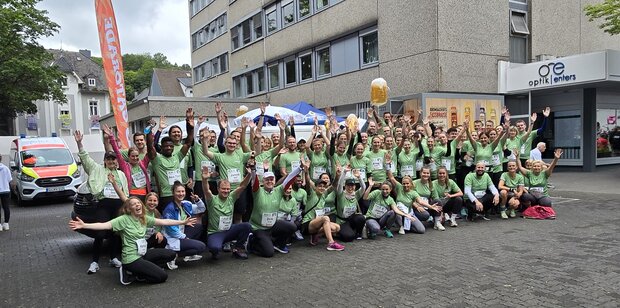 Gruppenselfie von etwa 50 Personen in grünen T-Shirts, die vor einem Gebäude stehen. Viele heben die Arme in die Luft und lächeln. 