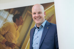 Mann mit Glatze in einem blauen Anzug und hellblauer Hemd steht vor einer Wand. Er lächelt in die Kamera.