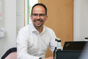 Ein Mann sitzt an einem Tisch vor einem Laptop, trägt ein weißes Hemd und lächelt in die Kamera. 