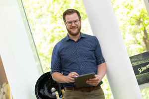 Mann mit blonden Haaren und trägt ein blaues Hemd. Er steht in einem hellen Raum und hält ein Tablet in der Hand und lächelt in die Kamera.
