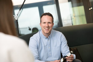 Mann mit kurz geschnittenen Haaren trägt ein hellblaues Hemd und sitzt auf einer dunkelgrünen Couch. Er lächelt in die Kamera.