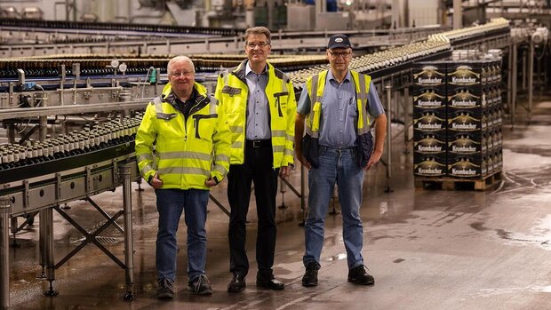 Drei Personen in gelben Warnjacken stehen in einer Fabrikhalle. Im Hintergrund sind Flaschen auf einem Förderband und Kisten zu sehen.