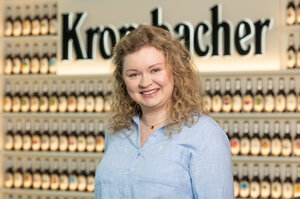 Frau mit lockigem, blondem Haar trägt ein hellblaues Hemd und lächelt in die Kamera.