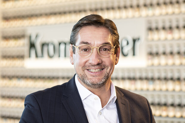 Mann mit braunen Haaren, trägt eine Brille und Dreitagebart. Dazu trägt er einen dunkelblauen Anzug. Er lächelt in die Kamera.