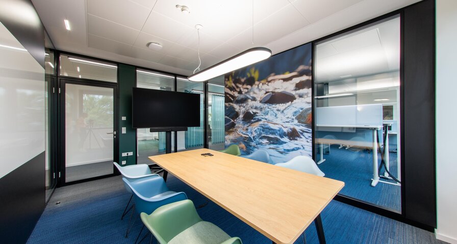 Besprechungsraum mit einem langen Holztisch und fünf Stühlen in verschiedenen Farben. An der Wand ein Bildschirm.