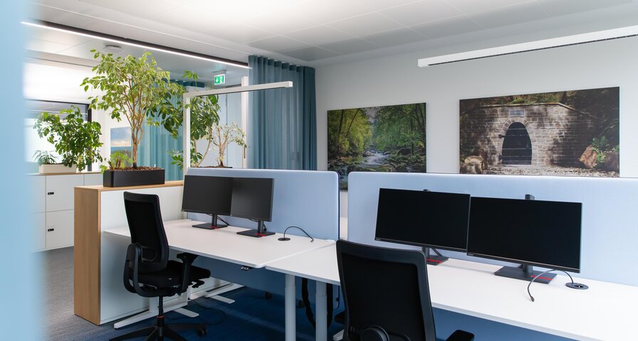 Büro mit zwei Schreibtischen, jeweils mit zwei Monitoren. Im Hintergrund sind Pflanzen und zwei Bilder an der Wand zu sehen.