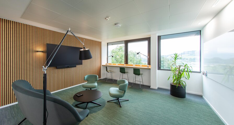 Moderne Büroumgebung mit Holzvertäfelung, einem großen Fernseher an der Wand, zwei grünen Sesseln, einem runden Tisch und einer Pflanze.