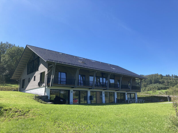 Zweistöckiges modernes Gebäude mit großen Fenstern und Balkon, umgeben von Wiese und Bäumen.