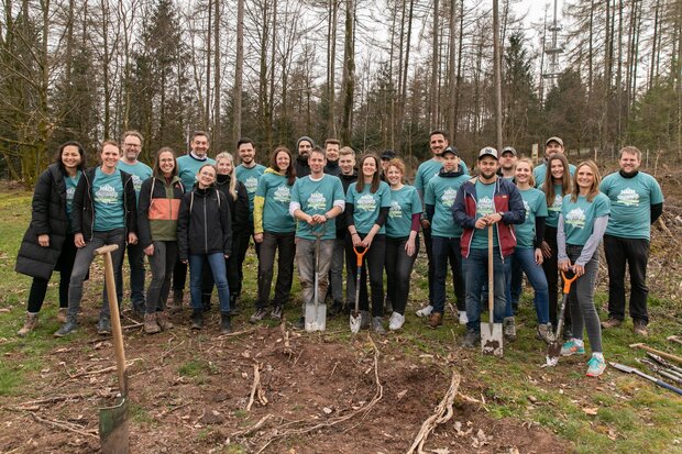 Gruppe von 20 Personen in blauen T-Shirts mit einem Baum-Motiv, stehen auf einer Waldlichtung. Einige halten Schaufeln und Spaten. Im Hintergrund sind Bäume sichtbar.