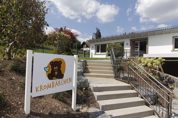 Ein Weg führt zu einem Gebäude mit einer weißen Fassade und einem grauen Dach. Links steht ein Schild mit der Aufschrift 'KROMBÄRCHEN' und einem Bären-Symbol. 