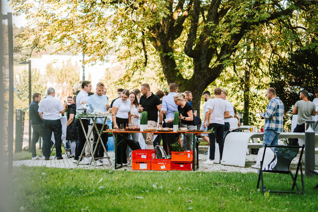 Eine Gruppe von Menschen steht in einem Garten. Im Vordergrund befindet sich ein Tisch mit Getränken und Snacks. 