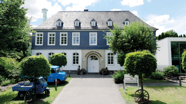 Krombacher Erlebniswelt, umgeben von Bäumen und Sträuchern. Vor dem Eingang stehen zwei große runde Bäume und ein blauer Lieferwagen.